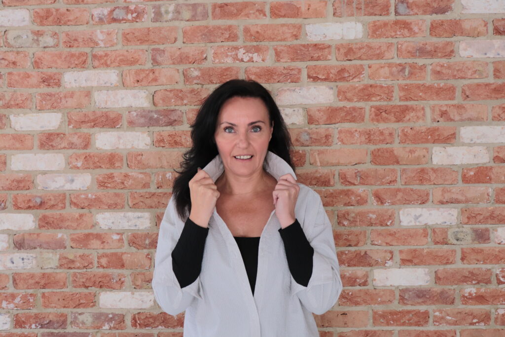 Michaela, transformational leadership coach, smiling confidently in front of a brick wall, representing empowerment and authenticity.